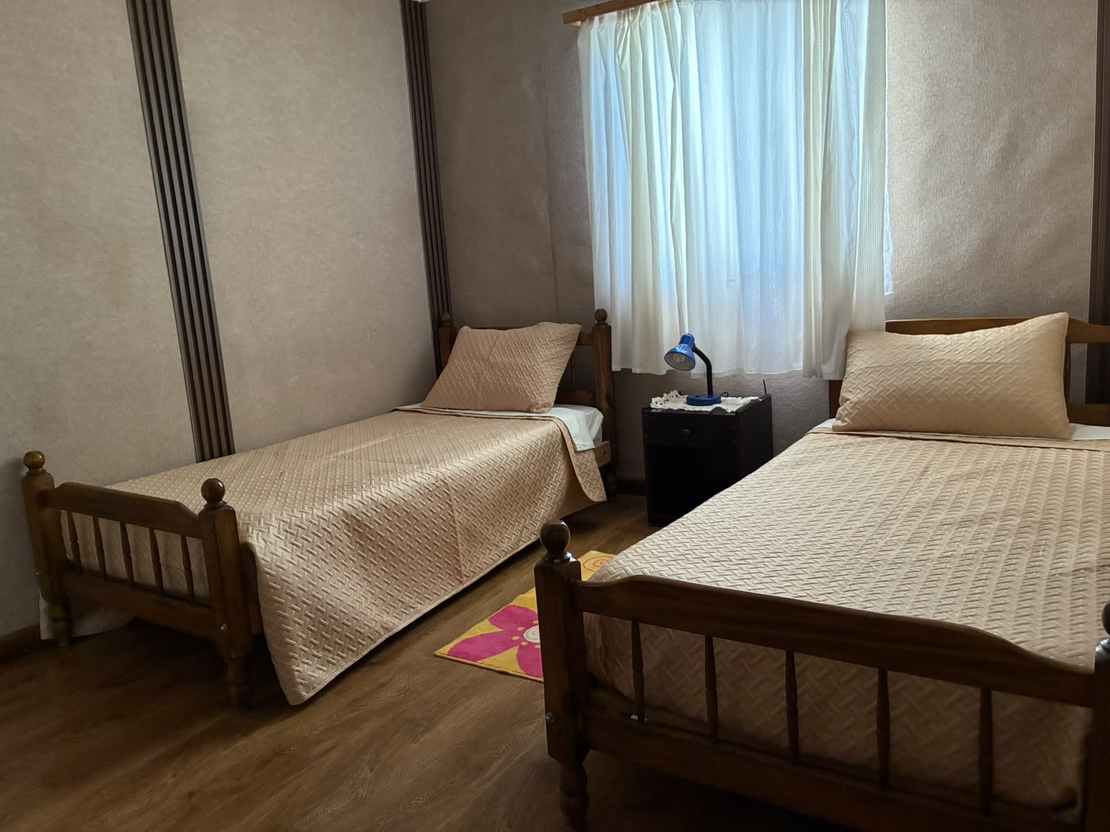Habitación con dos camas de madera, cubrecamas beige y una ventana con cortina blanca